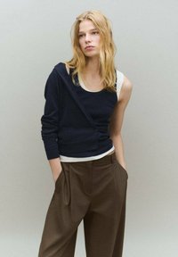 Cárdigan azul marino superpuesto a una camiseta blanca sin mangas, combinado con pantalones marrones de talle alto. Suave textura de punto y detalles de botones en el cárdigan.