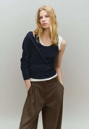 Cárdigan azul marino superpuesto a una camiseta blanca sin mangas, combinado con pantalones marrones de talle alto. Suave textura de punto y detalles de botones en el cárdigan.