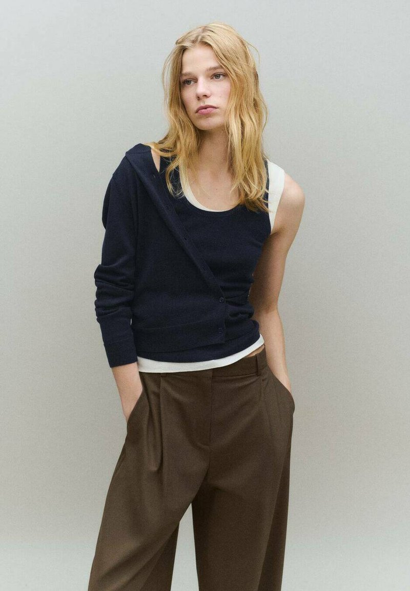 Cárdigan azul marino superpuesto a una camiseta blanca sin mangas, combinado con pantalones marrones de talle alto. Suave textura de punto y detalles de botones en el cárdigan.