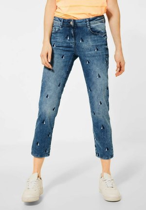 Personne portant un jean bleu court avec de petits motifs de voiliers, une chemise rayée orange et des baskets blanches, debout devant un fond blanc.