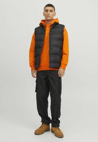 Sort puffervest over en orange hættebluse, parret med sorte cargo-bukser og brune støvler. Vestet har lynlåslukning og polstret design.