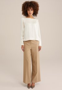 Top a maniche lunghe crema con scollatura a barca e texture delicata, abbinato a pantaloni beige a vita alta e gamba larga e tacchi grigi a punta.