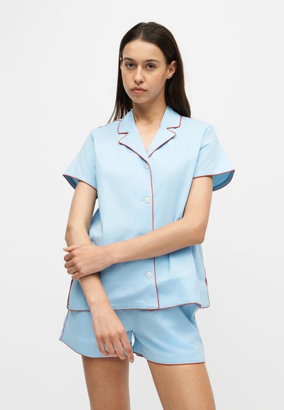 Jeune femme aux longs cheveux foncés portant un pyjama bleu clair à manches courtes avec boutons et bordure rouge, debout les bras croisés devant elle.