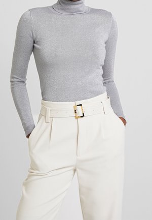 Femme portant un col roulé métallique argenté à manches longues, rentré dans un pantalon crème taille haute avec une ceinture assortie et une boucle dorée.