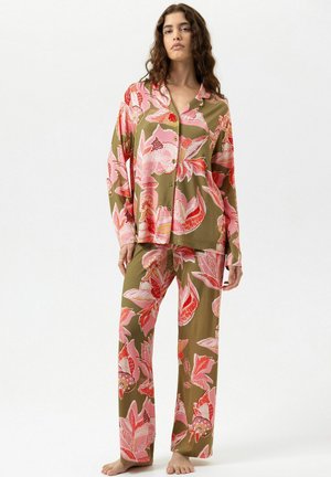 Vrouw die een olijfgroen hemdje met knopen en bijpassende wijde broek met grote roze en rode bloemdessin draagt, staat op blote voeten, neutrale achtergrond.