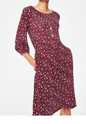 Kvinde iført en bordeauxrød kjole med blomsterprint, trekvartærmer, indsnøret talje, sidelommer og lagdelt gyldent vedhængshalskæde.