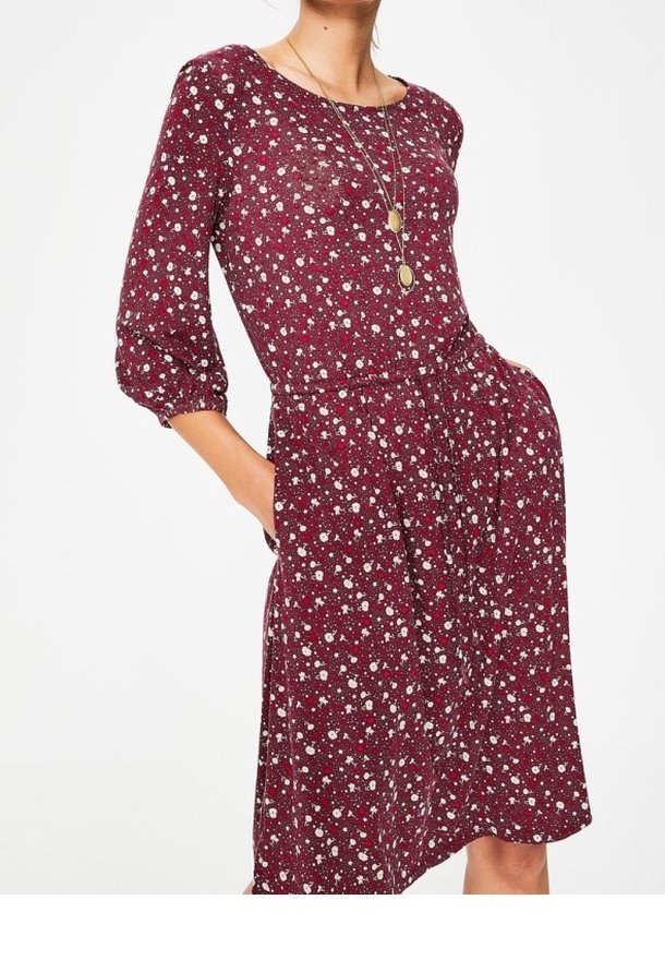 Femme portant une robe bordeaux à imprimé floral avec des manches trois-quarts, une taille cintrée, des poches latérales et un collier à pendentifs dorés superposés.