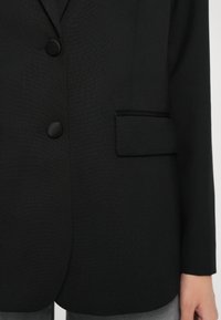 Blazer negru cu un material neted și texturat, două butoane mari și un singur buzunar cu detalii de margine contrastante.