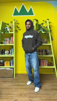 Felpa marrone con logo blu, jeans di denim azzurro chiaro e scarpe da ginnastica bianche. Sullo sfondo, pareti gialle e scaffali verdi con piante.