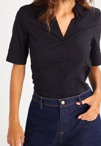 Sort kortærmet skjorte med knapper og krave, der har et tætsiddende design og glat tekstur, parret med mørkeblå denimbukser.