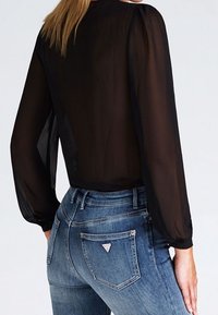 Top nero trasparente a maniche lunghe abbinato a jeans in denim aderenti di lavaggio medio, con logo a triangolo sulla tasca posteriore. Tessuto morbido e leggero.