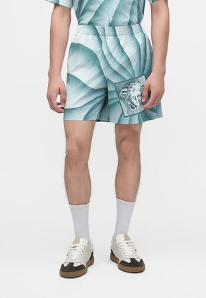 Mężczyzna noszący komplet szortów i koszuli w wzór zielononiebiesko-biały z nadrukiem głowy Meduzy, białe skarpetki oraz białe i czarne sneakersy.