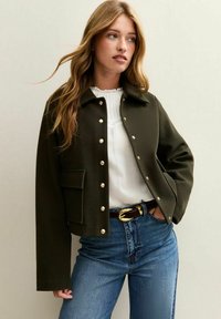 Vrouw met lang lichtbruin haar, gekleed in een olijfgroene jack met knopen over een witte blouse en blauwe spijkerbroek met een zwarte riem.