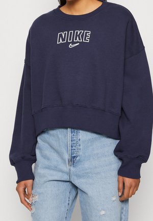 Sudadera corta azul marino de Nike con logo blanco, combinada con vaqueros rotos azul claro en una persona mostrada desde el cuello hasta las caderas.