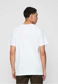 Camiseta de algodón blanca con mangas cortas y cuello redondo. Textura suave y corte relajado, mostrada desde atrás. Sin logotipos visibles.