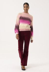 Strickpullover mit einem Farbverlauf von Pink zu Creme, kombiniert mit burgunderfarbenen Cordhosen im Schlag. Glatte Textur und gerippte Bündchen. Schwarze Schuhe.