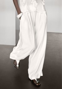 Witte, wijde broek met een gestructureerde stof en een gerimpelde tailleband, gecombineerd met zwarte sandalen met een zilveren accent bij de teen.