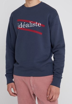 Person iført en marineblå sweatshirt med ordet "idéaliste" i hvid og understreget med to røde linjer, kombineret med lyserøde fløjlsbukser.