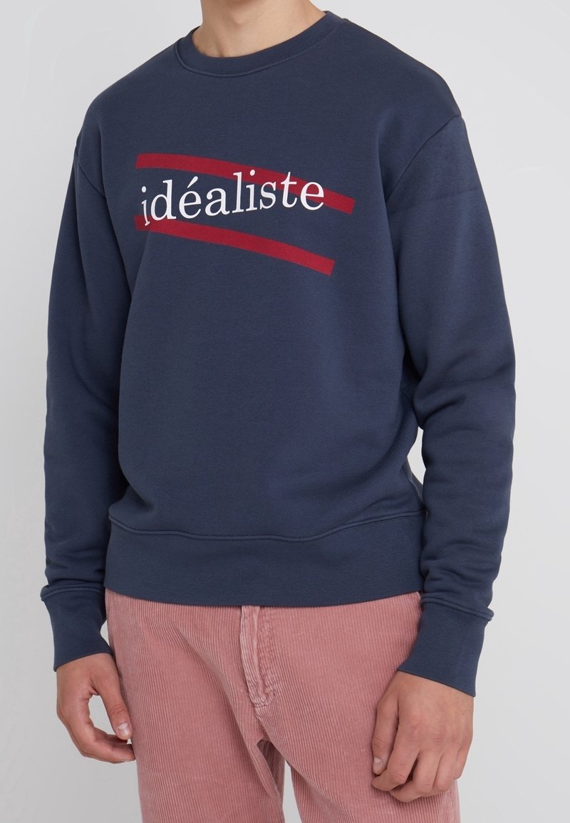 Personne portant un sweat-shirt bleu marine avec le mot « idéaliste » en blanc, souligné par deux lignes rouges, associé à un pantalon en velours côtelé rose.