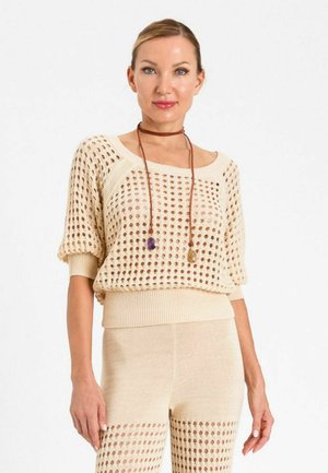 Donna che indossa top e pantaloni beige lavorati a maglia con ritagli circolari, accessoriati con una lunga collana marrone con pietre viola e gialle.