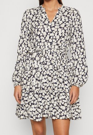 Frau trägt ein knielanges Kleid mit langen Ärmeln, dunkelblauem Hintergrund und weißem Blumenmuster, mit V-Ausschnitt und gebundenem Taillengürtel.