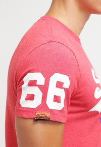 Maglietta rossa a maniche corte in cotone, con numero bianco "66" sulla manica sinistra e un'etichetta "SuperDry" vicino all'orlo.