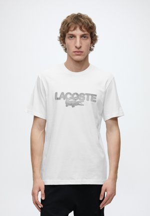 Ung mand med krøllet hår iført en hvid Lacoste t-shirt og sorte bukser, stående mod en ensartet hvid baggrund.