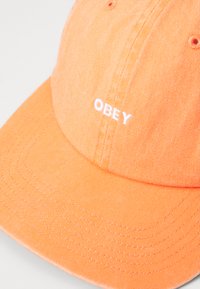 Narancssárga baseball sapka textúrált anyagból, fehér hímzett "OBEY" logóval és látható varrásokkal.