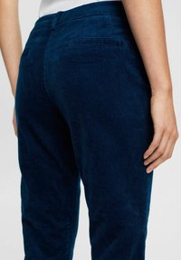 Gros plan d'une personne portant un pantalon en velours côtelé bleu foncé, montrant les détails des poches arrière et latérales avec une main droite visible.