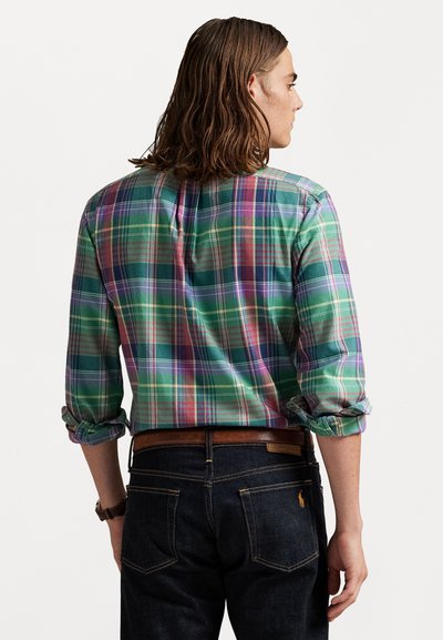Personne avec des cheveux châtains mi-longs portant une chemise à carreaux verte, violette et rose portée à l'intérieur d'un jean foncé avec une ceinture marron, de dos.