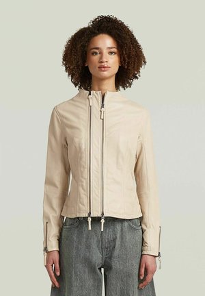 Donna con capelli ricci che indossa una giacca beige con cerniera e jeans grigi a vita alta, in piedi davanti a uno sfondo chiaro.