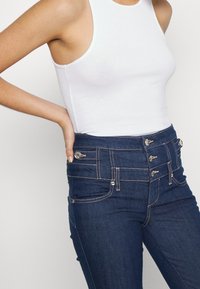 Mörkblå högmidjade jeans med flera knappdetaljer, kontraststygn och framfickor, kombinerat med en vit utanärmad topp.