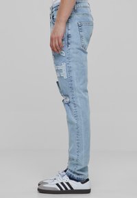 Jeans en denim bleu clair avec des détails déchirés, une coupe slim, cinq poches et une texture usée, associés à des baskets blanches avec des rayures noires.