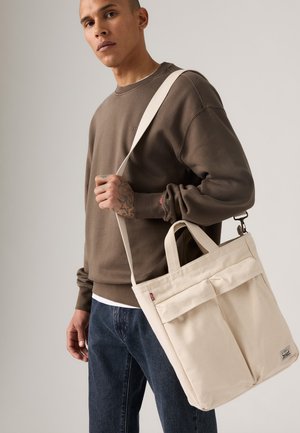 Bolsa de lona beige con dos bolsillos frontales y un asa superior, llevada al hombro. La modelo lleva un sudadera marrón y jeans oscuros.