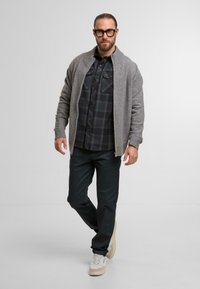 Šedý kardigán s rebrovanými manžetami, navlečený cez čiernu károvanú košeľu na gombíky. Tmavé nohavice a béžové tenisky dopĺňajú outfit. Jednoduchý, neformálny dizajn.