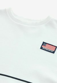 Bijela dukserica s rebrastim izrezom i crnim detaljima. Ima plavi pravokutni aplikaciju s američkom zastavom i tekstom "SUPERDRY".