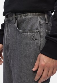 Jeans de denim gris con un ajuste relajado, que presentan un bolsillo frontal, un logo sutil y un acabado texturizado. Llevados con una camiseta de manga larga negra.