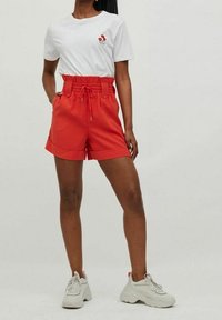 Pantalones cortos rojos con cintura de cordón y dobladillo doblado. Combinados con una camiseta blanca que presenta un pequeño diseño floral. Se llevan con deportivas blancas gruesas.