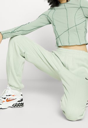 Person trägt hellgrünes, gesteppte langärmlige Oberteil und passende Jogginghosen, kniet und weiße und orangene Nike-Sneaker sind sichtbar.