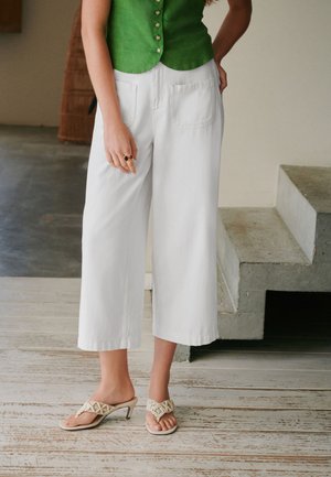 Kvinde iført grøn ærmeløs top, hvide vidde cropped bukser og beige hælede sandaler stående indendørs ved betonstrappen.