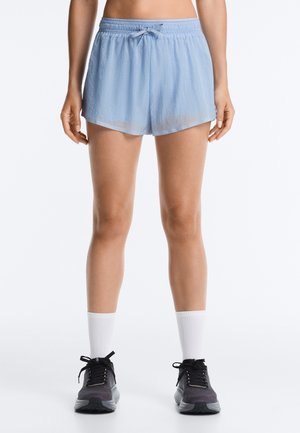 Vrouw draagt lichtblauwe gestructureerde korte broek, witte sportsokken en zwarte hardloopschoenen, staand tegen een effen witte achtergrond.