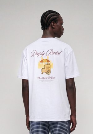Mand med flettet hår iført en hvid T-shirt med et billede af en hotdog-vogn og teksten "Deeply Rooted Brooklyn, New York" på ryggen.