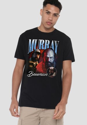 Junger Mann trägt ein schwarzes T-Shirt mit drei Porträts eines bärtigen Mannes und dem Text "Murray Bauman" auf der Vorderseite.