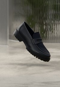 Μαύρα δερμάτινα μοκασίνια με υφασμάτινη υφή στο πάνω μέρος, στρογγυλή μύτη και χοντρή λαστιχένια σόλα, με διακοσμητική λουρίδα στη μπροστινή πλευρά.