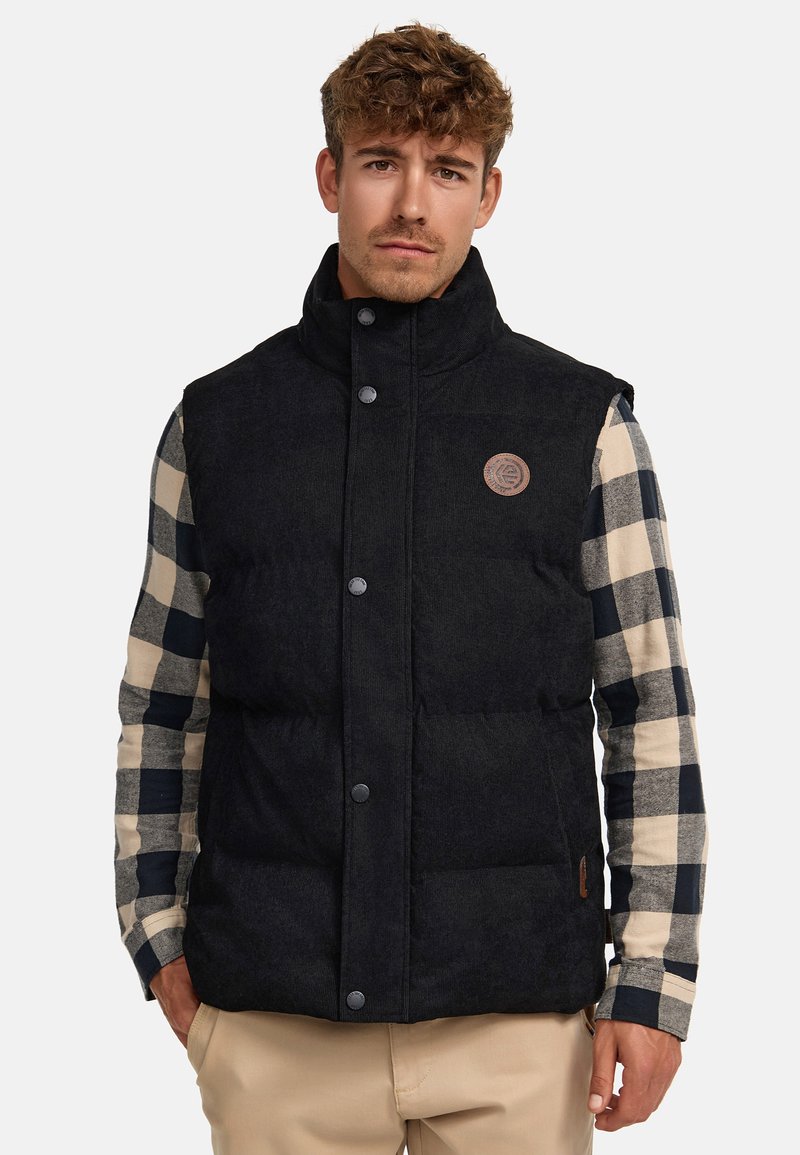 Gilet matelassé noir avec un col montant, des boutons-pression et une finition texturée. Présente un détail de logo et porté par-dessus une chemise à carreaux.