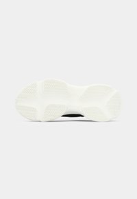 Sole of black sneaker showing thick white tread with zigzag patterns, against plain light gray background.