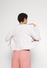 Biały, teksturowany kardigan z okrągłym dekoltem i dekoracyjnym wykończeniem. Stylizowany z różowymi spodniami, wykazuje luźny krój i otwarty tył.