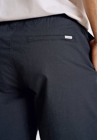 Mörkblå byxor med en bakficka som har en liten etikett. Tyget ser slätt ut med en lätt lyster och en resårmidja.