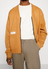 Homme portant un cardigan en laine moutarde sur un T-shirt blanc et un pantalon beige, avec une main dans la poche et une tâche visible sur la poche du cardigan.