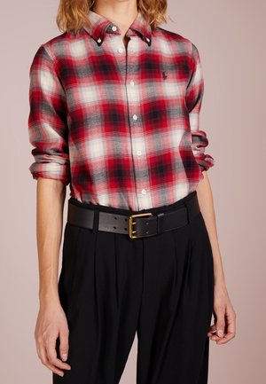 Person iført en rød, sort og hvid ternet skjorte med opkrogede ærmer, sorte bukser og et sort bælte med en guldlås.